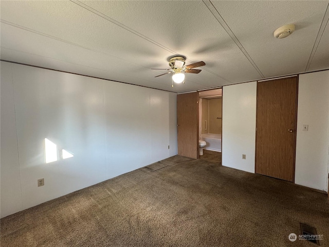 unfurnished bedroom with ceiling fan, ensuite bath, dark carpet, and a textured ceiling