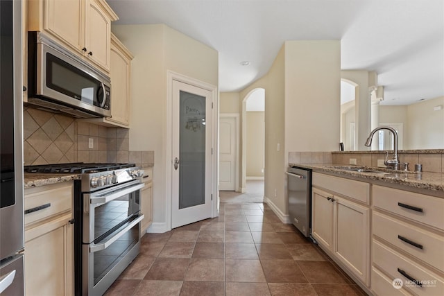 kitchen with sink, appliances with stainless steel finishes, light stone countertops, cream cabinets, and decorative backsplash