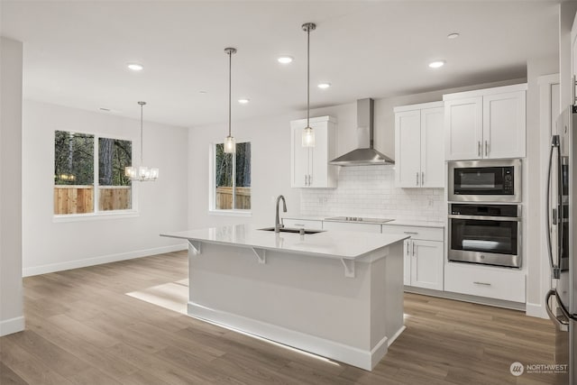 kitchen with sink, white cabinetry, an island with sink, stainless steel appliances, and wall chimney range hood