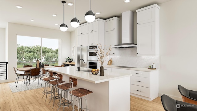 kitchen with an island with sink, white cabinets, hanging light fixtures, wall chimney range hood, and stainless steel double oven