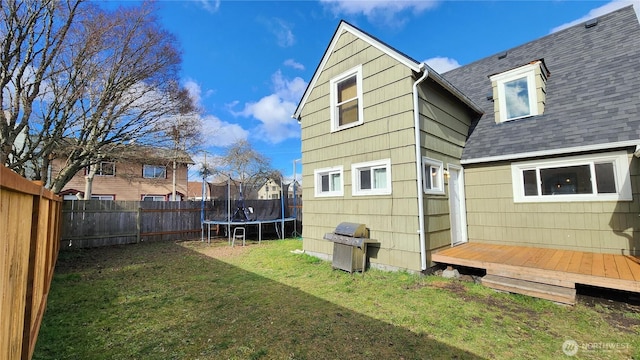 rear view of property featuring a deck, a fenced backyard, a yard, roof with shingles, and a trampoline