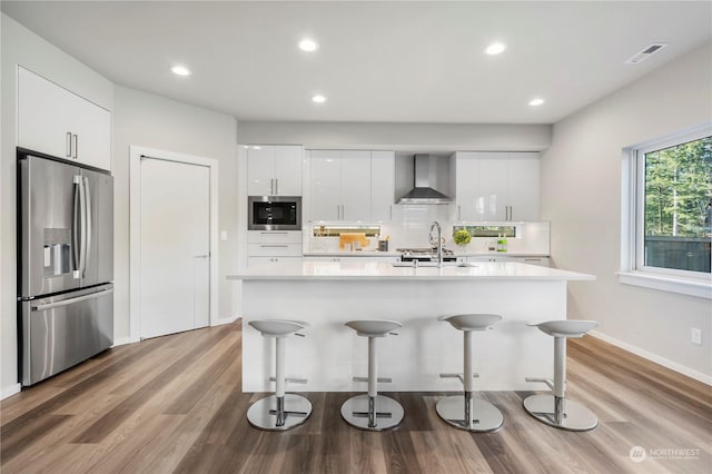 kitchen with stainless steel refrigerator with ice dispenser, built in microwave, a kitchen breakfast bar, wall chimney range hood, and white cabinets