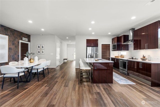 kitchen with appliances with stainless steel finishes, sink, a kitchen breakfast bar, wall chimney range hood, and a center island with sink