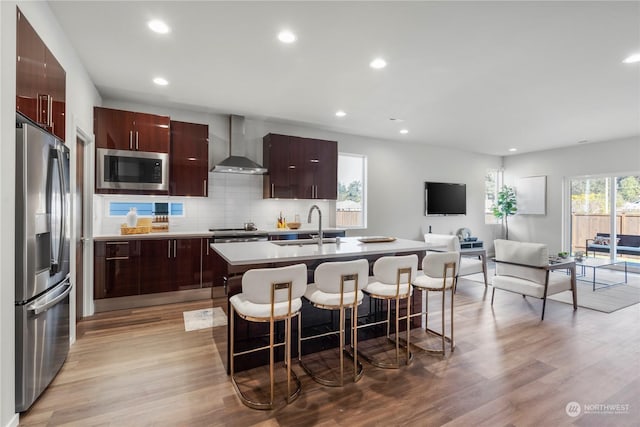 kitchen with wall chimney range hood, stainless steel fridge, a center island with sink, a kitchen bar, and built in microwave