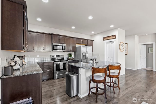 kitchen featuring dark wood-style flooring, appliances with stainless steel finishes, stone countertops, dark brown cabinetry, and a kitchen breakfast bar