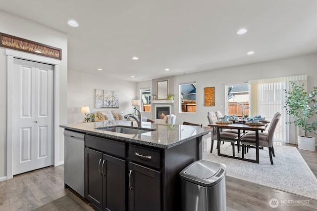 kitchen featuring stainless steel dishwasher, stone countertops, a glass covered fireplace, a sink, and an island with sink