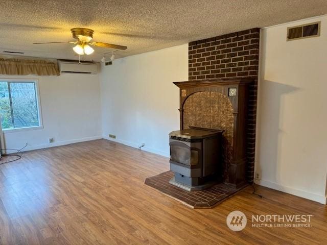 unfurnished living room featuring a textured ceiling, a wood stove, a wall unit AC, ceiling fan, and hardwood / wood-style floors