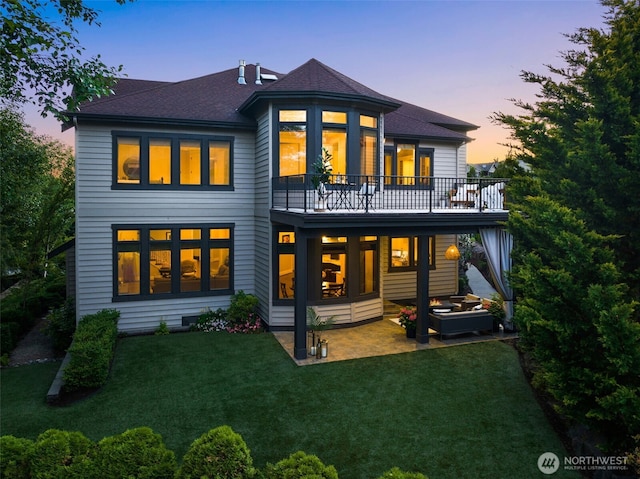 back of property at dusk featuring a shingled roof, a patio, a balcony, and a lawn