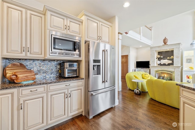 kitchen with appliances with stainless steel finishes, dark hardwood / wood-style floors, dark stone counters, cream cabinets, and backsplash