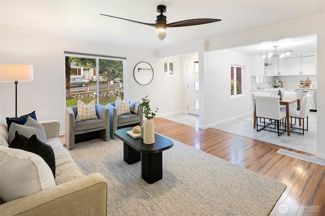 living room featuring light wood-style flooring, baseboards, crown molding, and ceiling fan with notable chandelier