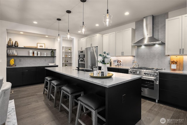 kitchen featuring high end appliances, light countertops, white cabinets, wall chimney range hood, and an island with sink