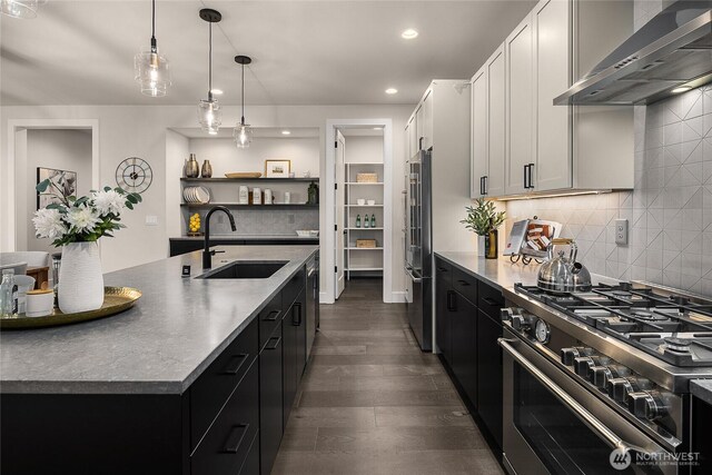 kitchen featuring premium appliances, a kitchen island with sink, white cabinets, a sink, and wall chimney range hood