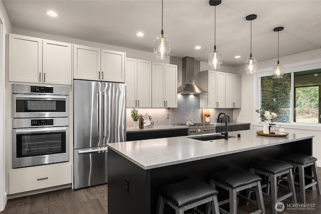 kitchen with pendant lighting, a center island with sink, stainless steel appliances, a sink, and wall chimney range hood