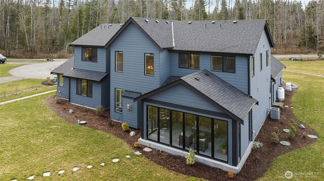 back of house with a shingled roof, a lawn, cooling unit, and a view of trees