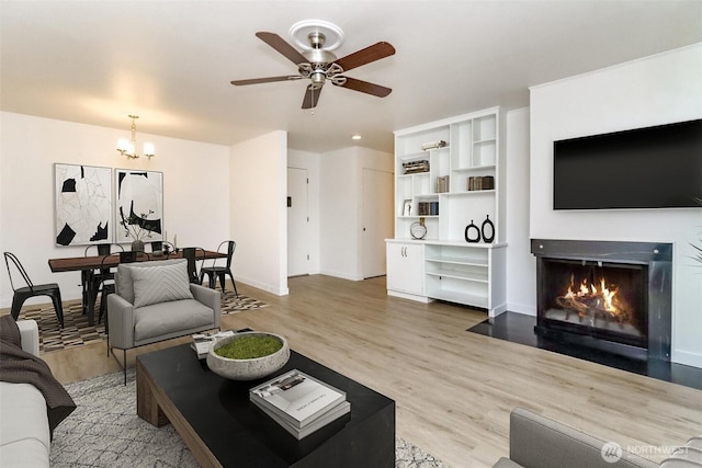 living area with light wood-type flooring, a fireplace with flush hearth, baseboards, and ceiling fan with notable chandelier