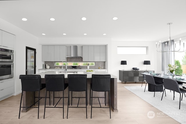 kitchen featuring a spacious island, a kitchen bar, stainless steel double oven, decorative backsplash, and wall chimney range hood