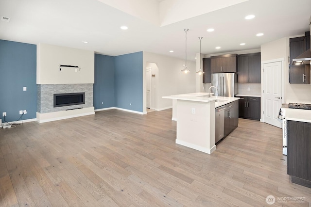 kitchen featuring appliances with stainless steel finishes, a stone fireplace, hanging light fixtures, an island with sink, and dark brown cabinetry