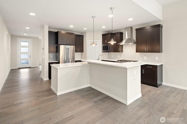 kitchen with a center island with sink, stainless steel appliances, dark brown cabinetry, decorative light fixtures, and wall chimney range hood