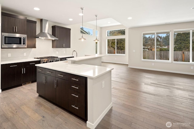 kitchen with appliances with stainless steel finishes, hanging light fixtures, wall chimney range hood, light wood-type flooring, and a kitchen island with sink