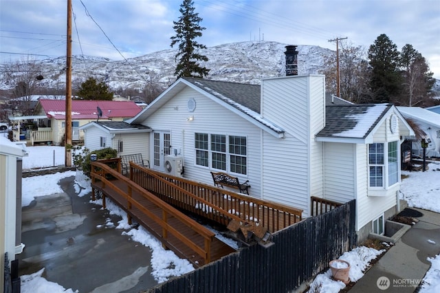 snow covered house with a wooden deck, a chimney, fence, and roof with shingles