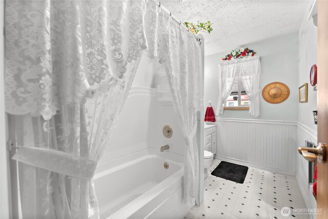 full bath with a wainscoted wall, shower / tub combo with curtain, toilet, a textured ceiling, and tile patterned floors