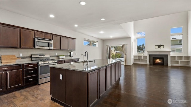 kitchen with appliances with stainless steel finishes, an island with sink, sink, dark brown cabinetry, and light stone counters