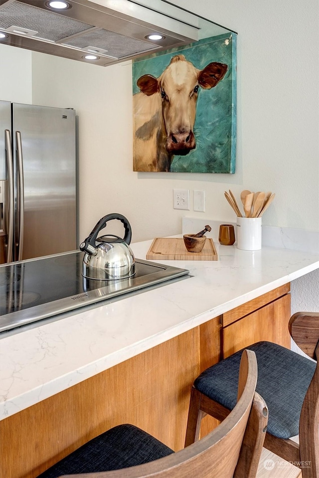 interior space featuring wall chimney range hood, stainless steel fridge, light stone countertops, and black stovetop