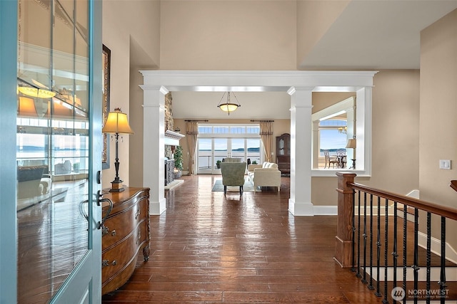 hallway with dark wood-style floors, baseboards, decorative columns, a towering ceiling, and an upstairs landing