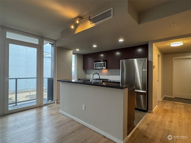 kitchen featuring appliances with stainless steel finishes, backsplash, a kitchen island with sink, dark brown cabinetry, and light wood-type flooring