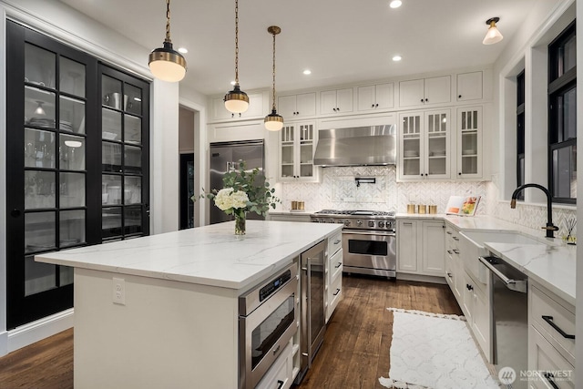 kitchen with built in appliances, a sink, a kitchen island, wall chimney range hood, and glass insert cabinets