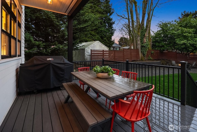 deck at dusk with outdoor dining area, a fenced backyard, a grill, a lawn, and a storage unit