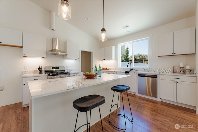 kitchen featuring a kitchen island, appliances with stainless steel finishes, white cabinetry, hanging light fixtures, and wall chimney range hood