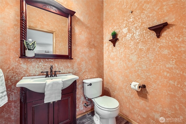 half bath with toilet, wood finished floors, vanity, and wallpapered walls