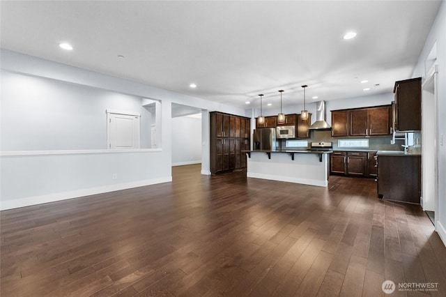 kitchen with a kitchen bar, dark wood-type flooring, stainless steel appliances, wall chimney exhaust hood, and dark brown cabinets