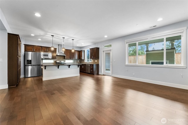 kitchen featuring a kitchen breakfast bar, dark wood finished floors, dark brown cabinetry, appliances with stainless steel finishes, and wall chimney range hood