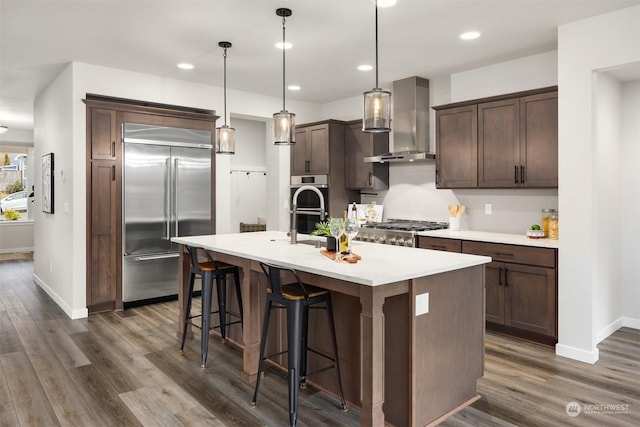 kitchen featuring dark brown cabinetry, a center island with sink, hanging light fixtures, stainless steel appliances, and wall chimney range hood