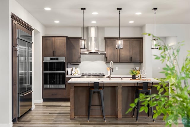 kitchen featuring pendant lighting, wall chimney range hood, stainless steel appliances, a kitchen breakfast bar, and dark brown cabinetry