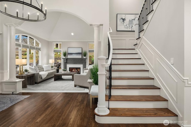 stairway featuring a decorative wall, wainscoting, wood finished floors, a lit fireplace, and ornate columns