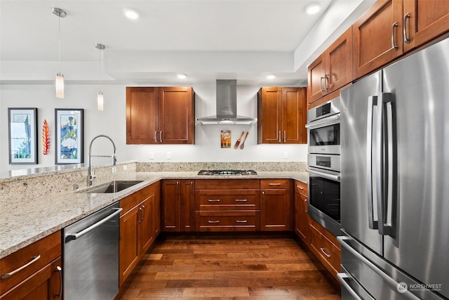 kitchen featuring brown cabinetry, wall chimney exhaust hood, appliances with stainless steel finishes, decorative light fixtures, and a sink