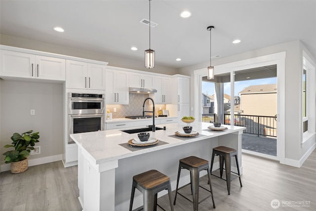 kitchen featuring a breakfast bar, pendant lighting, white cabinets, a kitchen island with sink, and stainless steel double oven