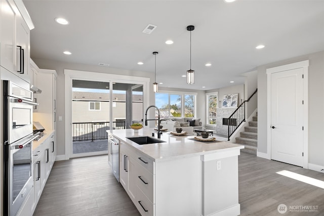 kitchen with sink, appliances with stainless steel finishes, a kitchen island with sink, white cabinetry, and decorative light fixtures