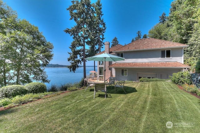 rear view of property with an attached garage, a lawn, a water view, and a chimney