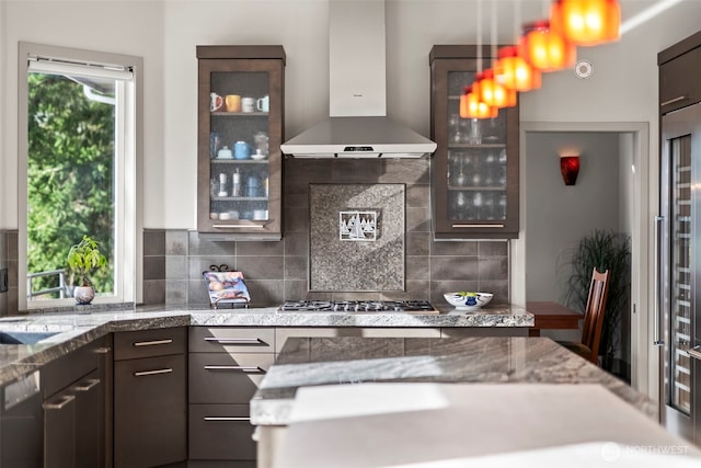 kitchen featuring dark brown cabinetry, stainless steel gas cooktop, dishwasher, wall chimney exhaust hood, and glass insert cabinets