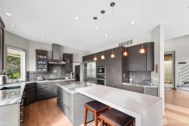 kitchen featuring wall chimney exhaust hood, glass insert cabinets, built in appliances, hanging light fixtures, and a sink