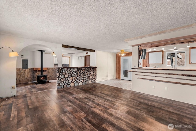 living area with ceiling fan, a textured ceiling, wood finished floors, electric panel, and a wood stove