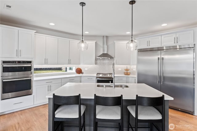 kitchen with wall chimney range hood, appliances with stainless steel finishes, white cabinets, a center island with sink, and decorative light fixtures