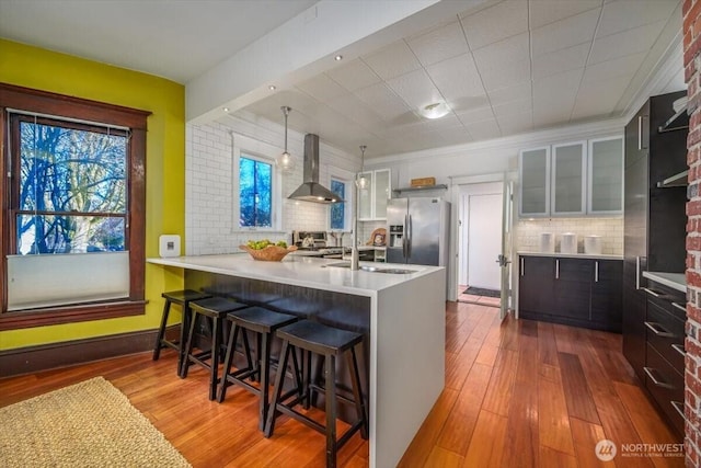 kitchen with wall chimney range hood, hardwood / wood-style flooring, a breakfast bar area, stainless steel fridge with ice dispenser, and kitchen peninsula