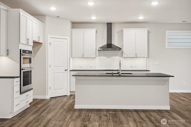 kitchen with double oven, white cabinetry, an island with sink, dark hardwood / wood-style flooring, and wall chimney exhaust hood