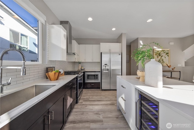 kitchen featuring wall chimney range hood, sink, appliances with stainless steel finishes, white cabinetry, and wine cooler