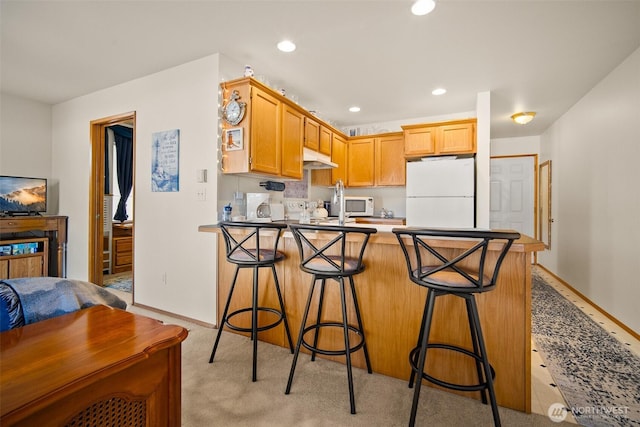 kitchen featuring a breakfast bar, light colored carpet, kitchen peninsula, light brown cabinets, and white appliances